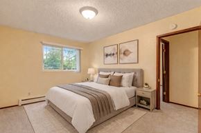 Anchorage apartment for rent - A serene Brighton Woods bedroom featuring a large window that frames green treetops and fills the room with daylight. A plush bed dressed in white linens and warm earth-tone pillows sits against a soft yellow wall, complemented by matching nightstands, soft carpet, and simple abstract artwork for a peaceful retreat.