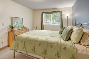 Anchorage apartment for rent - A charming Brighton Woods bedroom decorated in soft green tones with a neatly made bed featuring botanical-patterned bedding and layered pillows. A large window framed by matching curtains brings in natural light, while a light wood dresser with artwork and a tall floor lamp add warmth to the cozy space.