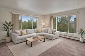 Anchorage apartment for rent - bright living room here at Castle featuring a large L-shaped sofa with soft neutral pillows, a wooden coffee table, and decorative plants adding warmth to the space. Oversized windows fill the room with natural light and offer leafy views, creating an inviting place to relax and unwind.