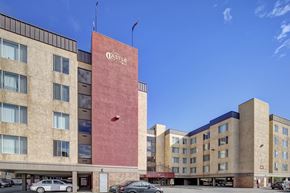 Anchorage apartment for rent - A bright exterior view here at Castle showing the multi-story beige and red building beneath a clear blue sky. Large windows line the façade, with the bold red central tower displaying the Castle name. Doorways, parking areas, and cars are visible at ground level, creating a welcoming first impression of the community.