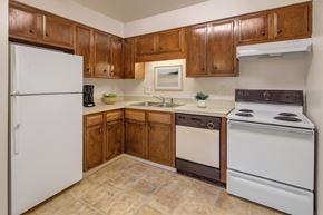 Anchorage apartment for rent - A warm kitchen here at Castle featuring rich wood cabinetry, a white refrigerator and stove, and a spacious L-shaped countertop with a double sink. Simple décor like a framed print, a coffee maker, and bowls of fruit adds inviting touches, while soft lighting brightens the cozy layout.