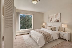 Anchorage apartment for rent - A serene bedroom here at Castle showcases a neatly made bed with white bedding, soft tan throws, and coordinating pillows set against two modern abstract prints. Plush carpet runs wall to wall while large windows bring in natural light and showcase leafy treetop views beside warm bedside lighting on matching nightstands.