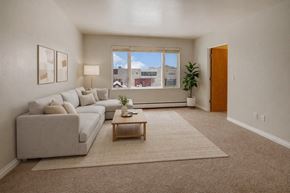 Anchorage apartment for rent - A bright living room here at Century Plaza featuring a large window that fills the space with natural light and frames a city view. A soft neutral sectional sits atop a textured rug beside a wooden coffee table, paired with modern wall art, a floor lamp, and a tall potted plant creating a calm, inviting atmosphere.