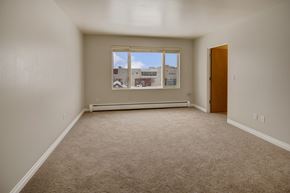 Anchorage apartment for rent - A bright living space here at Century Plaza showcasing an open layout with neutral wall tones and plush carpeting. A large picture window lets in natural light and offers a view of nearby buildings under a clear blue sky, while an adjoining doorway connects the room to the rest of the apartment.