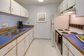Anchorage apartment for rent - A bright kitchen here at Chugach South featuring clean white cabinetry with wood trim, vivid blue countertops, and matching upper and lower storage on both sides of the room. Stainless steel double sinks, a classic electric stove, and a full-size refrigerator sit beneath soft wall lighting, creating a tidy and welcoming cooking space.
