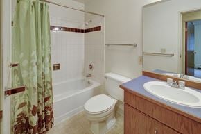 Anchorage apartment for rent - A bright bathroom here at Chugach South featuring a shower and bathtub combo with a green patterned curtain, white tile walls with a decorative accent strip, a standard toilet, and a wood vanity with a blue countertop and sink beneath a large mirror.