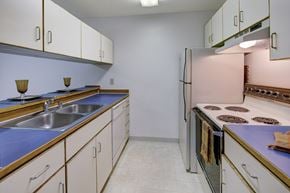Anchorage apartment for rent - A bright kitchen here at Chugach South featuring clean white cabinetry with wood trim, vivid blue countertops, and matching upper and lower storage on both sides of the room. Stainless steel double sinks, a classic electric stove, and a full-size refrigerator sit beneath soft wall lighting, creating a tidy and welcoming cooking space.