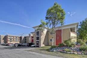 Anchorage apartment for rent - A sunny exterior view of City View Apartment Homes showcasing modern multi-story buildings with bold red and tan accents, balconies, and well-kept landscaping framed by rocks, shrubs, and trees. A clear blue sky, parked cars, and the welcoming entrance sign create a bright and inviting first impression of the community.