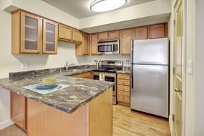Anchorage apartment for rent - A spacious kitchen here at City View featuring wood cabinetry, stainless steel appliances, a large marbled countertop peninsula with place setting, modern lighting, and wood-style flooring.