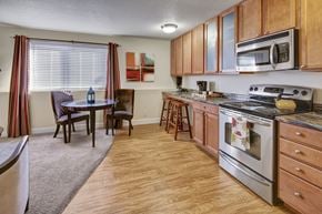 Anchorage apartment for rent - A bright open kitchen and dining space here at City View featuring warm wood cabinets, stainless steel appliances, a glass-top dining table with two chairs, striped curtains framing a large window, and wood-style flooring flowing into carpeted living space.