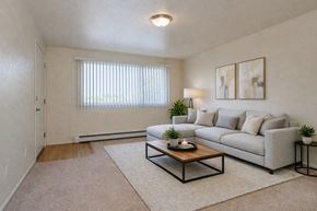 Anchorage apartment for rent - A bright living room here at Club at Eagle Pointe featuring a light gray sectional sofa with neutral pillows, a wooden coffee table on a soft area rug, and modern abstract wall art. A wide window with vertical blinds brings in natural light, creating an open, comfortable space ideal for everyday living.