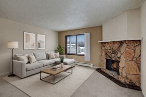 Anchorage apartment for rent - A cozy living room here at Conifer Grove featuring a light gray sectional sofa with neutral pillows, a simple wooden coffee table, and a soft area rug that defines the seating space. A large window brings in natural light, while a striking stone fireplace adds warmth and character to the room.