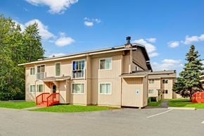 Anchorage apartment for rent - A bright exterior view here at Conifer Grove featuring a well-kept apartment building with neutral-toned siding, white-trimmed windows, and small private balconies, complemented by red entry railings, paved walkways, nearby parking, and surrounding greenery under a clear blue sky.
