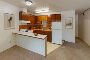 Anchorage apartment for rent - A functional kitchen here at Country Lane featuring warm wood cabinetry, white appliances, and an open breakfast bar overlooking the living area, with neutral flooring, simple wall art, and bright overhead lighting creating a clean and practical apartment layout.