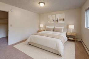 Anchorage apartment for rent - A bright bedroom here at Country Lane featuring a queen-sized bed with white bedding and neutral accent pillows, matching nightstands with lamps, soft carpeting, an area rug under the bed, abstract wall art, and an open closet providing simple storage and a calm, comfortable layout.