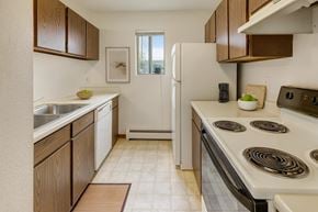 Anchorage apartment for rent - A bright kitchen here at Deer Park featuring warm wood cabinetry, white countertops, a double sink, dishwasher, electric range, and refrigerator, with a compact galley layout, vinyl flooring, neutral wall art, and a window that brings in natural light.