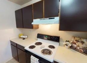 Anchorage apartment for rent - A functional kitchen here at Deer Park showcasing dark wood upper and lower cabinets, light countertops, an electric range with coil burners, a built-in vent hood, and simple decorative accents, with a clean, efficient layout designed for everyday cooking and convenient meal preparation.
