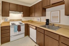 Anchorage apartment for rent - A functional kitchen here at Driftwood showcasing warm wood cabinetry, ample countertop space, a stainless sink, white electric range with vent hood, built-in dishwasher, neutral wall art, and simple decor accents that create a clean, practical layout ideal for everyday cooking and meal prep.