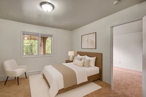 Anchorage apartment for rent - A calm bedroom here at Driftwood featuring soft carpet flooring, a spacious layout, a cozy bed with neutral bedding and accent pillows, a warm wood headboard, bedside table with lamp, natural light from the window, and leading into an adjacent room, creating a comfortable and inviting retreat.