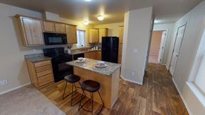 Anchorage apartment for rent - A warm, inviting kitchen here at Four Seasons featuring natural wood cabinetry, granite-style countertops, a central island with seating, black appliances, and wood-look flooring, with ceiling lighting, a nearby hallway for easy flow, and an open layout that connects seamlessly to the living space for everyday comfort.