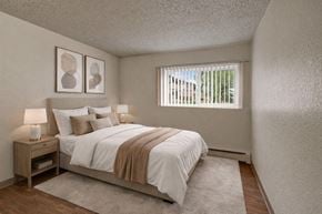 Anchorage apartment for rent - A warm and inviting bedroom here at The Greenbriar featuring a neatly made bed with crisp white bedding, layered neutral accent pillows, a soft beige throw blanket, matching nightstands with modern lamps, abstract wall art above the headboard, wood-style flooring, a plush area rug, and a window with vertical blinds providing natural light and a peaceful outdoor view.