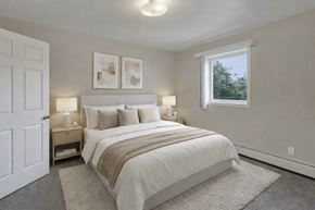 Anchorage apartment for rent - A bright bedroom here at La Maisonnette with beige carpet and soft neutral walls. The room features spacious bed with white bedding, neutral accent pillows, and a light throw blanket, flanked by two matching bedside tables with lamps. A window brings in natural light and offers a view of greenery outside, creating a calm and inviting atmosphere.