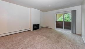 Anchorage apartment for rent - An open-concept and empty living room here at Ladera Villa, with beige carpet and neutral walls. A fireplace is set into the wall, and a sliding glass door opens to a private balcony with greenery outside, creating a welcoming atmosphere.