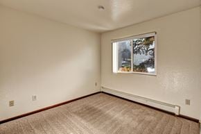 Anchorage apartment for rent - An open and empty bedroom space here at Ladera Villa, with a beige carpet and neutral walls. A window brings in natural light and views of trees outside, with baseboard heating along the wall.