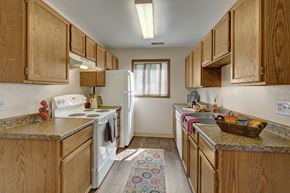 Anchorage apartment for rent - A kitchen here at Legacy West with light wood cabinets, beige countertops, and wood-look flooring. The space includes a white stove, white refrigerator, a double sink, and a window with blinds for natural light.