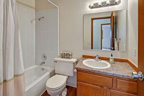 Anchorage apartment for rent - A bright, clean bathroom at Legacy West featuring a bathtub-shower combo with a white curtain and tile surround. A white toilet sits next to a wood vanity with a sink, mirror, and overhead lighting.