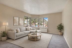 Juneau apartment for rent - A bright, cozy living room here at Mendenhall-Tower with a light gray sectional sofa, a round wooden coffee table, and a soft neutral area rug. A wall of large corner windows brings in beautiful natural light and scenic neighborhood views for an airy, inviting atmosphere.