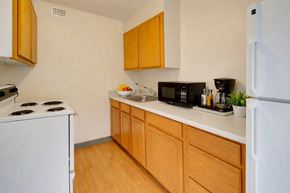 Juneau apartment for rent - A compact, functional kitchen here at Mendenhall-Tower with light wood cabinets, clean white countertops, and warm wood-style flooring. A white stove and refrigerator sit alongside a stainless steel sink, with a cozy coffee station and small greenery adding a welcoming touch.