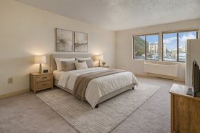 Juneau apartment for rent - A bright, cozy bedroom here at Mendenhall-Tower with soft neutral carpet, a comfortable bed dressed in white linens, and warm bedside lighting. A large window brings in natural light and scenic city views, creating an airy and relaxing atmosphere.