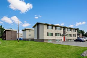 Anchorage apartment for rent - A three-story Montclair apartment building with a neutral exterior, a bright red entry door, and open green lawn space under a sunny blue sky.