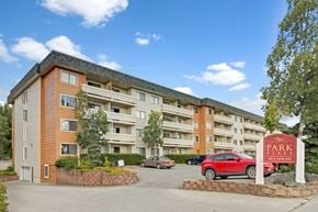 Anchorage apartment for rent - A welcoming exterior view of the Park Plaza apartment community with a multi-story building featuring private balconies, mature landscaping, and convenient on-site parking beneath a bright blue sky.