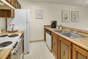 Anchorage apartment for rent - A clean, cozy galley kitchen with warm wood cabinets and butcher-block style countertops, featuring a white refrigerator, white stove, double stainless sink, and simple wall decor with framed botanical prints.