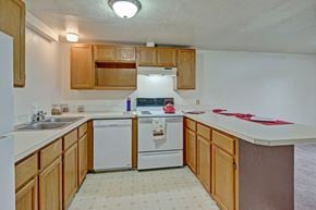 Anchorage apartment for rent - A spacious kitchen here at Ptarmigan Meadows with light wood cabinets and bright white countertops, featuring a double stainless steel sink, a white dishwasher and stove, and a breakfast bar that opens into the living area.