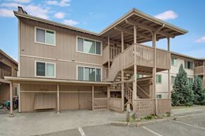 Anchorage apartment for rent - The exterior of a Rancho Tudor apartment building with beige siding, shared stairwell balconies, and nearby parking spaces under a bright blue sky.