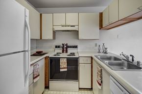 Anchorage apartment for rent - A compact kitchen here at Silver Ridge with cream-colored cabinets, white countertops, and warm wood trim for a cozy, classic feel. A white refrigerator and electric stove sit beneath a range hood, while a double stainless steel sink and dishwasher add everyday convenience.