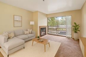Anchorage apartment for rent - A bright living room here at Susitna Ridge with a cozy light-gray sectional sofa, a wooden coffee table, and soft carpet flooring, featuring a corner fireplace and a sliding glass door that opens to a private balcony with natural views.
