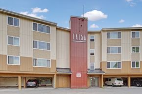 Anchorage apartment for rent - A bold exterior view here at Taiga featuring a multi-story apartment building with a central Taiga Apartment Homes sign, large windows, and covered parking below under a bright blue sky.