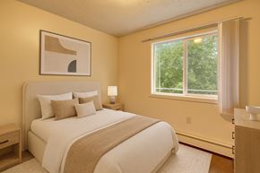 Anchorage apartment for rent - A warm, inviting bedroom here at Taiga with soft neutral walls, a cozy bed with layered pillows, a bedside table and lamp, and a large window with vertical blinds overlooking leafy green views.