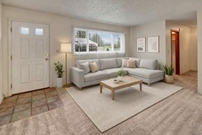 Anchorage apartment for rent - A bright living room here at Terrace on the Lake with a light gray sectional sofa, neutral throw pillows, a wooden coffee table on a beige rug, and a large window with vertical blinds that brings in natural light and outdoor views.