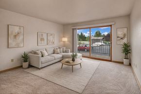 Anchorage apartment for rent - A bright living room at The Alaskan featuring plush carpet, a modern sectional sofa, and a round coffee table on a neutral rug. Soft artwork and plants add warmth, while a large sliding glass door opens to a balcony with views of the parking area and nearby greenery.