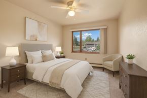 Anchorage apartment for rent - A bright bedroom at The Alaskan with soft carpeting and warm neutral walls. The space features a neatly made bed with matching bedding, two bedside tables with lamps, and a dresser. A large window brings in natural light and offers views of the surrounding neighborhood.