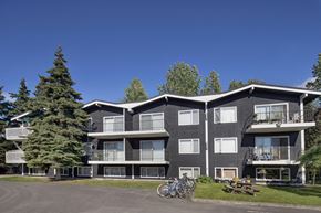 Anchorage apartment for rent - A three-story exterior view of The Alaskan with dark siding, white trim, and private balconies on each level. Mature evergreens and open lawn space create a natural setting, with a picnic table and bicycles adding to the community-friendly atmosphere under a bright blue sky.