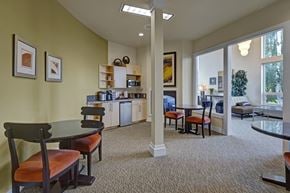 Anchorage apartment for rent - A cozy resident café area at The Alpine featuring small round tables with cushioned seating, a beverage station with a microwave, coffee maker, and mini fridge, and open shelving for supplies. Warm earth tones, framed artwork, and carpeted floors create a relaxed space next to the building’s lobby.