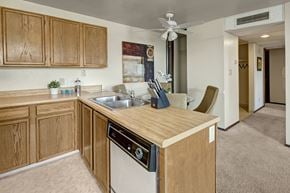 Anchorage apartment for rent - A kitchen at The Alpine featuring wood cabinets, laminate countertops, and a double sink with a dishwasher below. The space opens to a dining nook with upholstered chairs and a ceiling fan overhead. Neutral carpet and a hallway leading to other rooms complete the open layout.