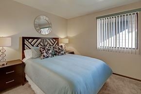Anchorage apartment for rent - A cozy bedroom at The Alpine featuring a neatly made bed with blue bedding and patterned accent pillows. Matching bedside tables with lamps sit on each side of the headboard, and a window with vertical blinds brings in natural light. A round wall mirror adds a stylish touch above the bed.