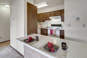 Anchorage apartment for rent - A functional kitchen here at The Continental featuring dark wood cabinetry, white appliances including a stove and dishwasher, laminate countertops with a breakfast bar, overhead lighting, and simple place settings that highlight the open layout connecting to the living area.